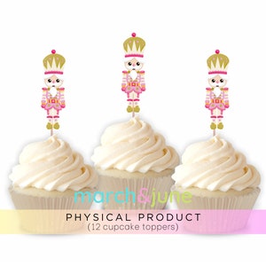 May include: Twelve pink and gold nutcracker cupcake toppers. The nutcrackers are wearing pink and gold outfits and have white faces. The toppers are on white frosted cupcakes.