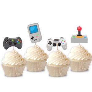 May include: Four cupcakes with different video game themed toppers. The toppers are a black and green game controller, a gray and white game controller, a gray handheld game console, and a gray joystick with a red button.