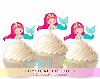 May include: Twelve cupcake toppers featuring a pink-haired mermaid with a blue tail and a gold crown. The mermaids are holding their hands together in front of them. The toppers are on white frosted cupcakes.