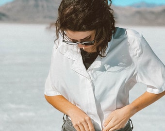 May include: A woman wearing a white button-down shirt with a black lace trim and dark gray denim shorts. She is adjusting the waistband of her shorts.