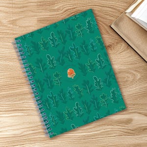 Peut inclure: Un cahier à spirales avec une couverture turquoise ornée d'un motif de feuilles vertes et d'une petite illustration de gland. Le cahier est ouvert sur une surface en bois, avec un second livre visible en arrière-plan.
