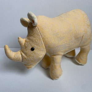 May include: A plush rhino toy in a light yellow fabric with a subtle floral pattern. The rhino has small black eyes, a horn, and soft, cream-colored accents on its ears and feet. The toy is designed to stand on four legs.