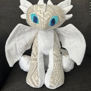 May include: White plush dragon with light brown patterned fabric on its body and wings. The dragon has blue eyes and a white, fluffy tail.
