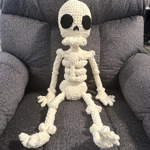 May include: A handmade, off-white crochet skeleton doll with black eye sockets. The plush figure has articulated limbs and a detailed rib cage, sitting on a gray upholstered chair. The soft, textured yarn gives it a whimsical, Halloween-themed appearance.
