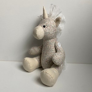 May include: A plush unicorn toy with a white lace trim on its head. The unicorn is made of a beige fabric with a floral pattern and has a white button on its arm.