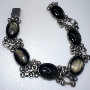 May include: A silver-toned bracelet featuring oval-shaped black gemstones with gold flecks. The bracelet has a series of connected links and a clasp closure. The gemstones are set in a decorative silver metal setting.