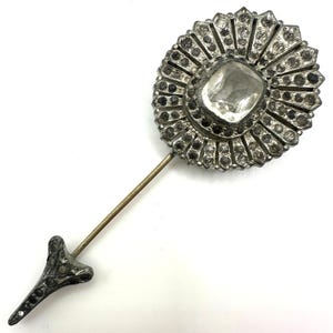 Puede incluir: Un broche de sombrero plateado vintage con una gran piedra central transparente de talla cuadrada, rodeada de piedras más pequeñas y transparentes. El broche tiene un extremo decorativo y puntiagudo y un eje largo y recto. El diseño es ornamentado y detallado.