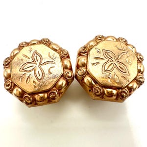 May include: A pair of gold-toned metal earrings with an octagonal shape. Each earring features a central engraved four-petaled floral motif surrounded by a decorative border of raised, rounded beads and small spiral scrollwork details.