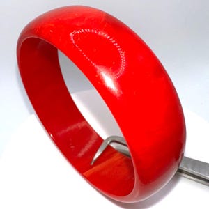 May include: A vibrant red bangle bracelet. The smooth, glossy surface reflects light, highlighting its rounded shape. The bracelet is a solid, bold color, and appears to be made of a hard material.