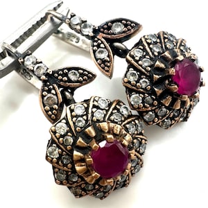 May include: A pair of sterling silver drop earrings featuring a 925 hallmark, each with a central round deep pink stone surrounded by small clear stones in a floral pattern, set in a darkened metal finish with leaf-shaped accents.