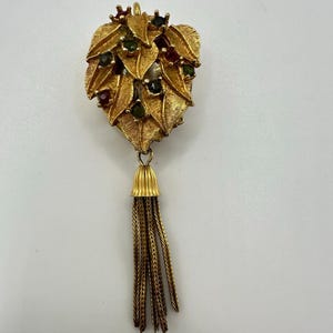 May include: A gold-toned vintage brooch in the shape of a leaf, adorned with small red and green gemstones. The brooch features detailed leaf veins and a tassel of fine chains hanging from a bell-shaped base. A classic piece of jewelry.
