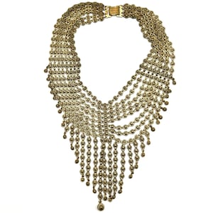 May include: A gold-toned statement necklace with multiple strands of delicate chains and dangling beaded accents. The necklace features a decorative clasp and a cascading design, creating a bold and elegant look. The necklace is suitable for special occasions.