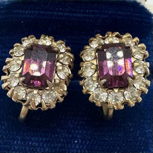 May include: A pair of vintage clip-on earrings. Each earring features a rectangular, deep purple gemstone surrounded by small, clear crystals. The setting is gold-toned metal. The earrings are displayed against a dark blue velvet background.