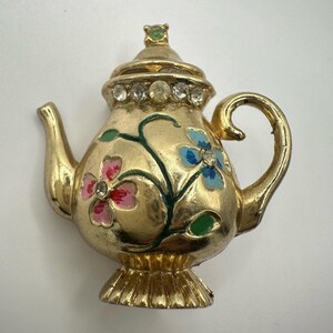 May include: A gold teapot brooch with a floral design. The teapot has a lid with rhinestones and a handle. The flowers are painted in pink, red, blue, and green.