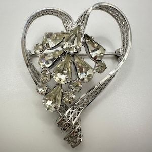 May include: Silver-toned brooch in the shape of a heart, adorned with clear, teardrop-shaped and round rhinestones. The heart's outline features an openwork design, with a decorative swirl at the bottom. A pin secures the brooch.