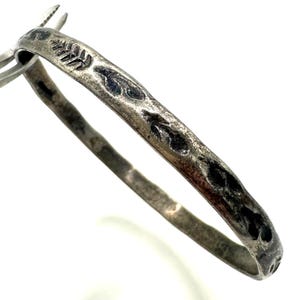 Peut inclure: Un bracelet jonc en métal argenté avec une finition vieillie et oxydée. La surface extérieure présente une série de motifs en relief rappelant des feuilles, assombris dans les zones en creux, créant un aspect texturé et organique.