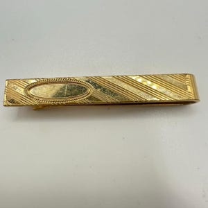 May include: A gold-toned tie clip with an oval center and diagonal striped design. The clip has a hinged clasp and is made of a shiny metal. The tie clip is a classic accessory.