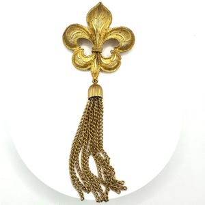 Peut inclure: Broche dorée représentant une fleur de lys avec une gland de chaînes. La broche présente une surface texturée et des détails complexes. Le gland est suspendu à un capuchon décoratif, ajoutant une touche d'élégance.