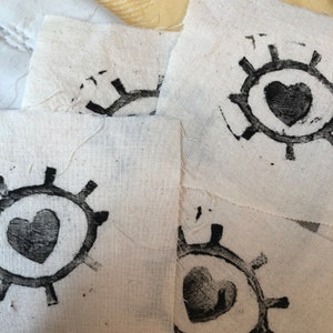 May include: Four white fabric squares with black ink stamps of a stylized eye with a heart in the center. The eye is surrounded by a circle with spokes radiating out from the center.
