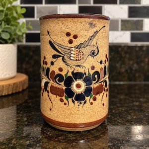 May include: A cylindrical ceramic mug with a speckled tan base featuring a hand-painted design of a bird perched above a large dark blue flower. The artwork includes intricate black line details, green leaves, and brown accents around the rim.