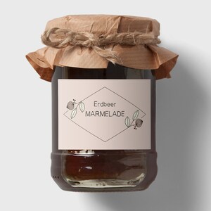 Könnte beinhalten: Ein Glas mit braunem Papierdeckel und einer Schnur darum gebunden. Das Glas enthält eine dunkelrote Marmelade. Das Etikett auf dem Glas lautet "Erdbeer Marmelade" in schwarzer Schrift mit einer stilisierten Illustration eines Erdbeerzweigs.