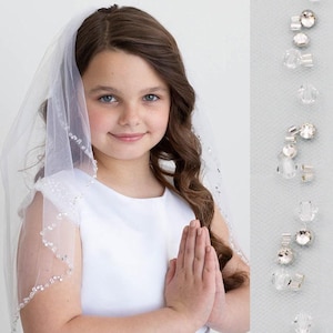 May include: A young person wearing a white dress and a sheer veil with beaded trim. The veil is adorned with small, sparkling beads. The dress has a simple, elegant design. The person is smiling and has their hands clasped together.