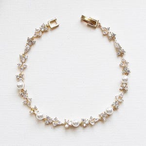 May include: A gold-toned bracelet featuring a delicate chain set with alternating clear, marquise-cut crystals and small, round white pearls. The piece includes a secure metal clasp closure, creating a circular design with a repeating floral-inspired pattern.