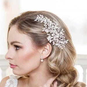 May include: A silver-toned hair comb featuring a floral-inspired arrangement of clear crystals and small white pearls. The decorative piece is secured into the side of wavy blonde hair, showcasing a detailed metal structure with leaf-like accents.