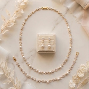 May include: A gold-toned jewelry set featuring a necklace, bracelet, and earrings. The necklace and bracelet are adorned with pearls and sparkling accents. The earrings are pearl drop earrings. The set is displayed on a neutral background with decorative elements.