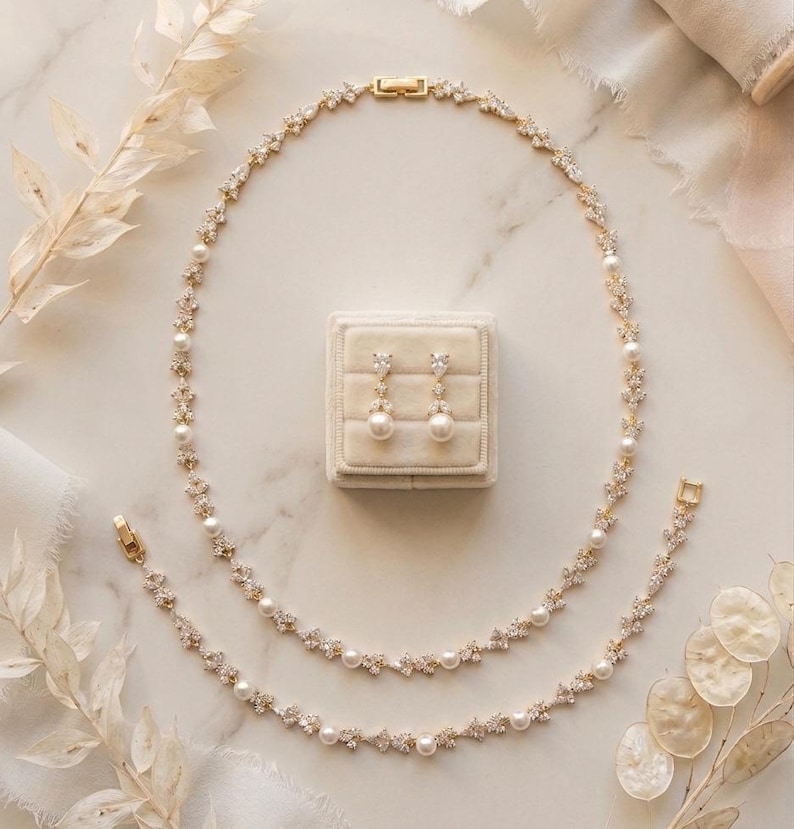 May include: A gold-toned jewelry set featuring a necklace, bracelet, and earrings. The necklace and bracelet are adorned with pearls and sparkling accents. The earrings are pearl drop earrings. The set is displayed on a neutral background with decorative elements.