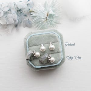 A collection of pearl earrings and a box of earrings is displayed on a white background, with a white flower and the words "Pierced Clip-Ons" written above.
