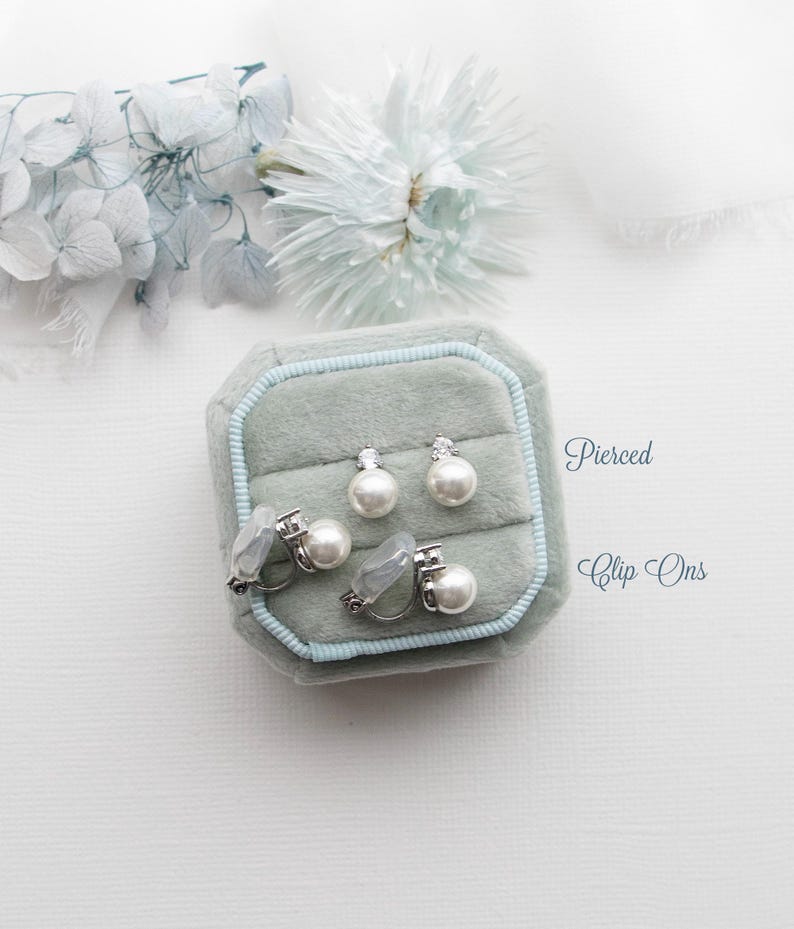 A collection of pearl earrings and a box of earrings is displayed on a white background, with a white flower and the words "Pierced Clip-Ons" written above.