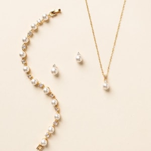 May include: A gold-toned jewelry set consisting of a bracelet, a necklace, and two stud earrings. The bracelet features alternating white pearls and clear crystals. The necklace has a thin gold chain with a single pearl and crystal pendant.