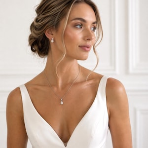 A woman in a white dress with her hair styled in an updo, wearing a necklace with a pendant.