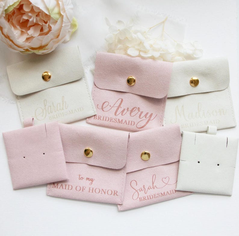 a collection of pink and white purses or bags, each with a personalized name printed on it, likely for bridesmaids. There is also a large pink flower in the background, adding a touch of elegance to the scene.