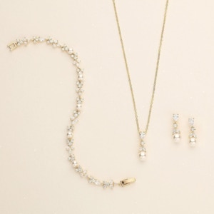 Puede incluir: Un conjunto de joyas dorado que incluye una pulsera, un collar y pendientes. La pulsera y los pendientes tienen un diseño floral con piedras transparentes y perlas. El collar tiene un colgante con una perla y piedras transparentes.