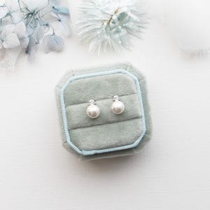 A pair of pearl earrings is displayed in a light blue velvet box, surrounded by a white background with a delicate flower arrangement.