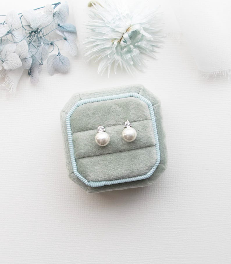 A pair of pearl earrings is displayed in a light blue velvet box, surrounded by a white background with a delicate flower arrangement.