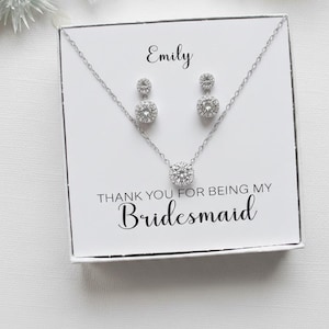 May include: A silver-toned necklace and matching drop earring set featuring round clear stones in a halo setting. The jewellery is presented in a white gift box with the printed text Emily and THANK YOU FOR BEING MY Bridesmaid in black ink.