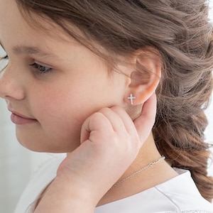 Peut inclure: Gros plan sur une oreille ornée d'une petite boucle d'oreille en forme de croix argentée. La boucle d'oreille est sertie de petites pierres claires. La personne porte un haut blanc et un collier argenté.