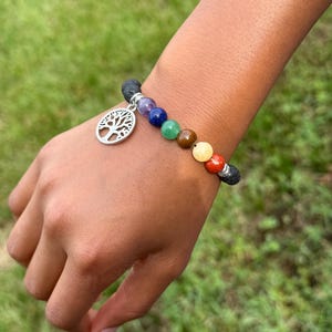 May include: A beaded bracelet featuring chakra colors and a silver tree of life charm. The bracelet includes a variety of colorful beads, including red, orange, yellow, green, blue, purple, and black lava beads. The bracelet is approximately 7 inches in circumference.