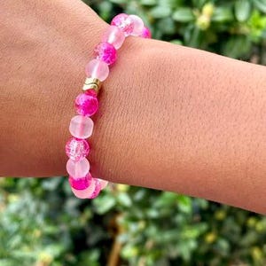 Puede incluir: Una pulsera de cuentas rosas con una mezcla de cuentas transparentes, opacas y texturizadas. La pulsera presenta un espaciador de color dorado y se usa en una muñeca. El fondo es follaje verde borroso.