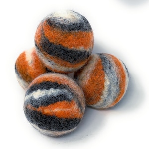 May include: A cluster of five felted wool balls. Each ball is round and features a striped pattern in orange, gray, white, and black. The balls are arranged on a white background. These are likely dryer balls.