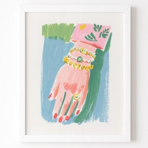 May include: A framed art print featuring a stylized illustration of a hand with red-painted fingernails, wearing three beaded bracelets in yellow, white, and green, and a gold ring with a circular stone, set against a blue and green background.