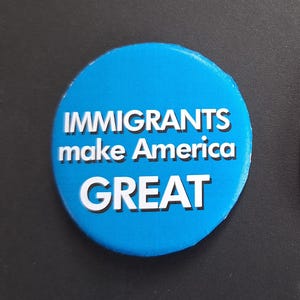 May include: A round, blue button with the white text "IMMIGRANTS make America GREAT". The button is a solid, bright blue color with white text. The text is in a bold, sans-serif font.