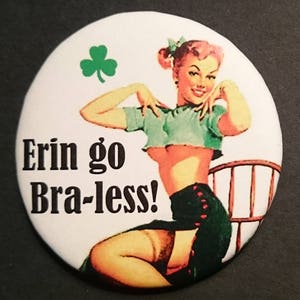 May include: A white button with a green shamrock in the top right corner. A woman in a green crop top and a black skirt is sitting on a wooden chair. The text "Erin go Bra-less!" is printed in black on the button.