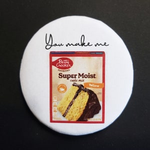 May include: A white round sticker with the text "You make me" printed in black. The sticker features a box of Betty Crocker Super Moist Yellow Cake Mix with a picture of a yellow cake with chocolate frosting.