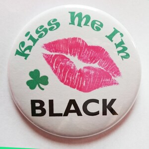 May include: A white button with green text that says "Kiss me I'm Black" and a pink lipstick print. There is a green shamrock in the corner.
