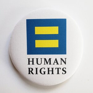 May include: A white button with a blue square containing two yellow horizontal bars. The text "HUMAN RIGHTS" is printed below the square.