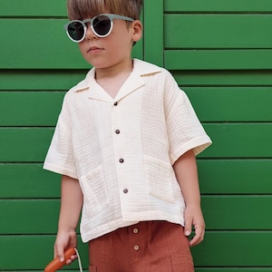May include: A child wearing a cream-colored short-sleeved shirt and rust-colored pants. The child is also wearing sunglasses and holding a blue and orange toy. The background is a green wooden door.